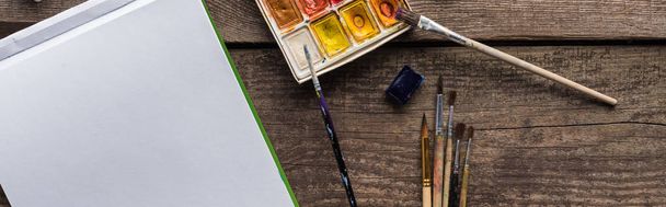 bovenaanzicht van kleurrijk verf palet, penselen en blanco schetsblok op houten oppervlak, panoramische foto - Foto, afbeelding