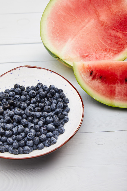 cut ripe red watermelon with seeds near plate with blueberries on wooden white table isolated on grey - Фото, зображення