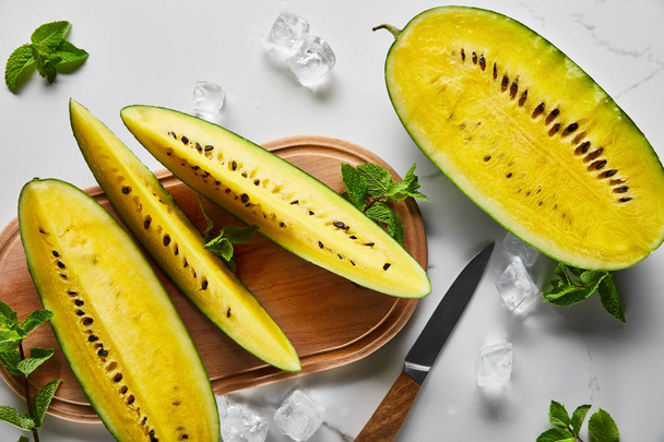 top view of cut delicious exotic yellow watermelon with seeds on wooden chopping board with knife, mint and ice cubes on marble surface - Photo, Image
