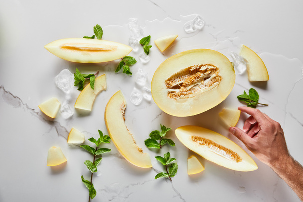 Draufsicht auf geschnittene köstliche Melone auf Marmoroberfläche mit Minze und Eis in der Nähe von Menschenhand - Foto, Bild