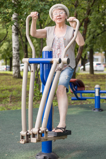 Μια ευτυχισμένη ηλικιωμένη γυναίκα οδηγεί έναν δραστήριο τρόπο ζωής, εξασκώντας σε εξομοιωτές στο πάρκο της πόλης το καλοκαίρι. - Φωτογραφία, εικόνα