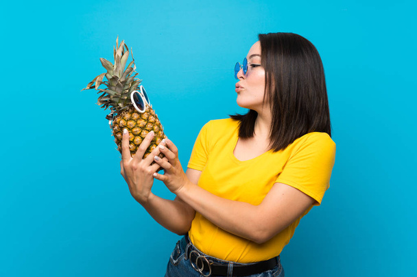 Молодая мексиканка на изолированном синем фоне держит ананас в солнечных очках
 - Фото, изображение