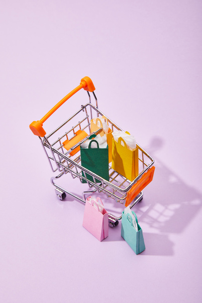 few small paper bags near toy cart with colorful shopping bags on violet background - Photo, Image