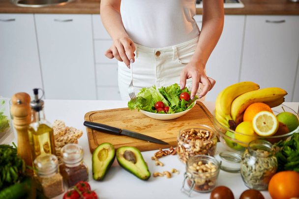 Молодая счастливая женщина готовит вкусный салат на красивой кухне с зелеными свежими ингредиентами в помещении. Концепция здорового питания и диеты. Потеря веса
 - Фото, изображение