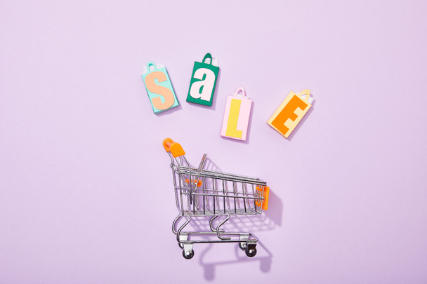 top view of colorful toy bags with sale lettering near shopping cart on violet - Foto, Bild