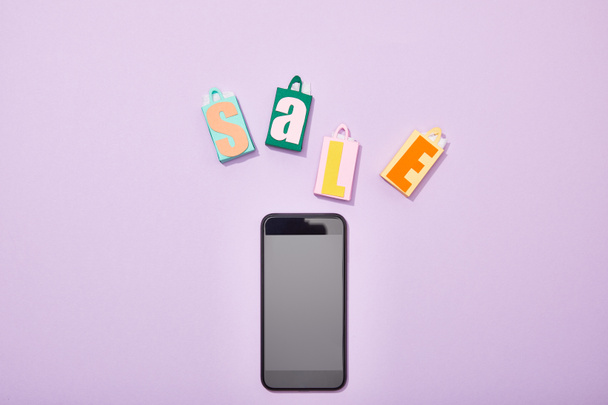 top view of colorful paper bags with sale lettering near smartphone with blank screen on violet  - Foto, afbeelding