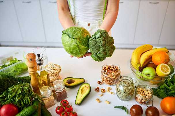 Молодая счастливая женщина держит брокколи и капусту на красивой кухне с зелеными свежими ингредиентами в помещении. Концепция здорового питания и диеты. Потеря веса
 - Фото, изображение