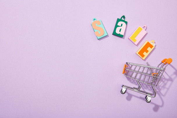 Draufsicht auf bunte Einkaufstüten mit Verkaufsaufschrift, die in einen Spielzeugwagen auf violett fallen  - Foto, Bild