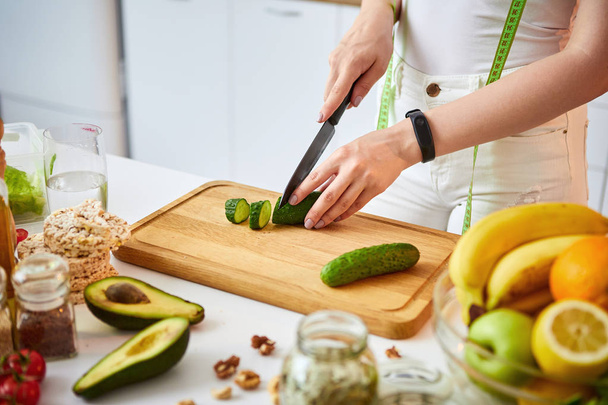 Jovem mulher feliz cortando pepino para fazer salada na bela cozinha com ingredientes frescos verdes dentro de casa. Alimentação saudável e conceito de dieta. Perder peso
 - Foto, Imagem