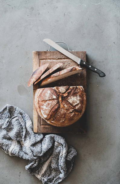 Flat-lay of freshly baked sourdough wholegrain bread loaf on rustic wooden board over grey concrete table background, top view - Foto, Imagen