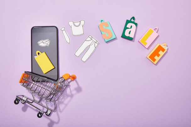 top view of smartphone with paper cut of clothes and yellow paper bag falling into toy cart near colorful shopping bags with sale lettering on violet  - Фото, зображення