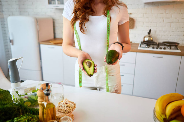 Young happy woman holding avocado for making salad in the beautiful kitchen with green fresh ingredients indoors. Healthy food and Dieting concept. Loosing Weight - Foto, Imagem