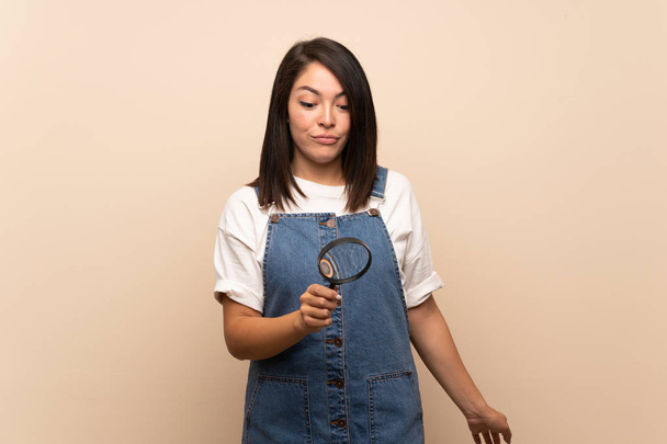 Young Mexican woman over isolated background holding a magnifying glass - Photo, image