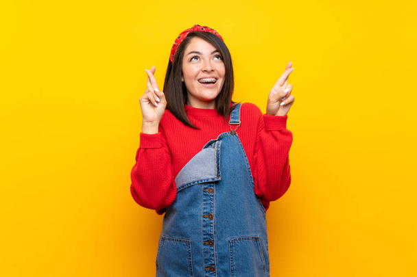 Young Mexican woman with overalls over yellow wall with fingers crossing and wishing the best - Фото, изображение