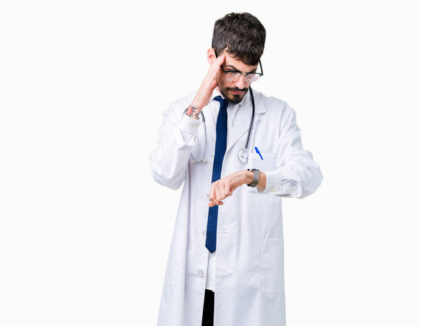 Jonge arts man dragen ziekenhuis vacht over geïsoleerde achtergrond kijken naar de tijd kijken bezorgd, bang om laat - Foto, afbeelding