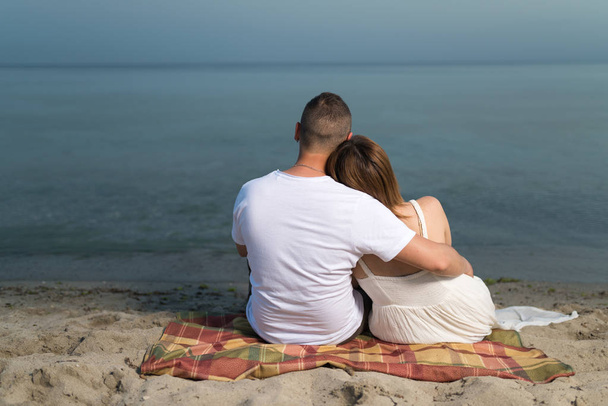Romantický portrét dvojice v zamilovaní objetí, sedícího na pláži při západu slunce. Na pozadí moře. - Fotografie, Obrázek