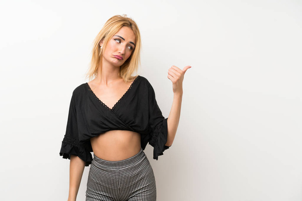 Young blonde woman over isolated white wall unhappy and pointing to the side - Photo, image