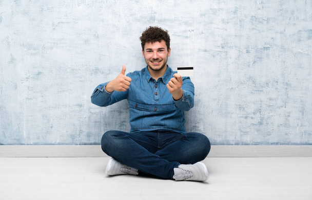 Młody mężczyzna siedzi na podłodze posiadając kartę kredytową - Zdjęcie, obraz