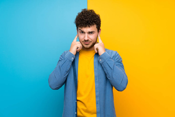 Muž se kudrnatými vlasy na barevné stěně frustrovaný a zakrývající uši - Fotografie, Obrázek