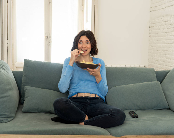 Happy woman on sofa with TV remote control ready to watch favorite movie of TV Show. Looking enthusiastic, making gestures of approval and eating chips. In people, technology and leisure concept. - Photo, Image