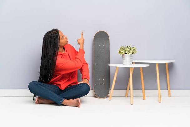 afrikanisch-amerikanische Skater Teenager Mädchen mit geflochtenen Haaren sitzt auf dem Boden und zeigt mit dem Zeigefinger nach hinten - Foto, Bild
