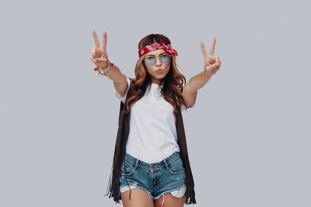 Attractive stylish young woman looking at camera and puckering while standing against grey background, gesturing peace with hands  - Foto, imagen