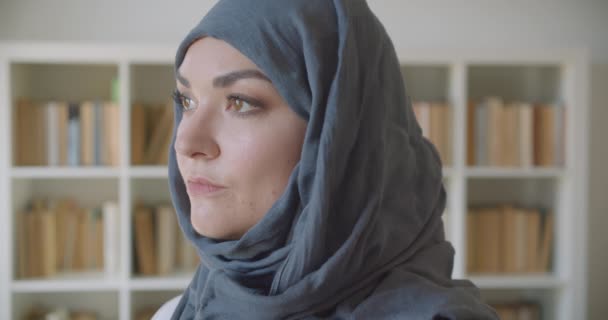 Closeup portrait of young muslim attractive businesswoman in hijab looking at camera in the library indoors with bookshelves on the background - Video, Çekim