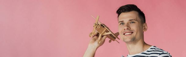 panoramic shot of smiling young man holding wooden toy plane and looking away isolated on pink - Photo, image