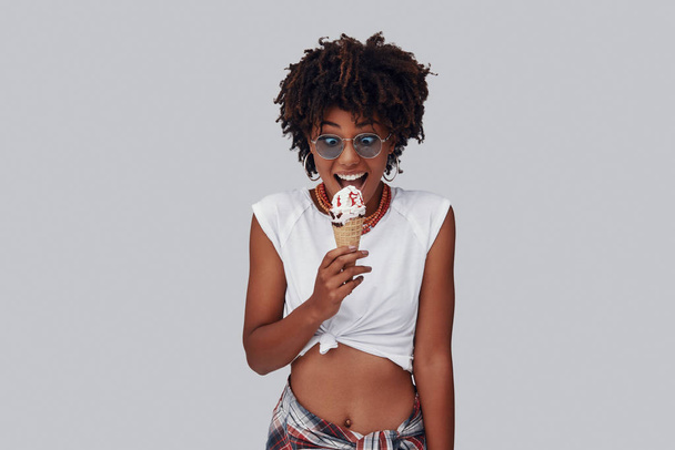 Attractive young African woman eating ice cream and smiling while standing against grey background - Foto, afbeelding