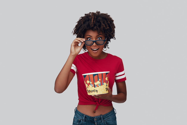 Attractive young African woman in 3D eyewear eating popcorn and looking at camera while standing against grey background - Foto, Imagem