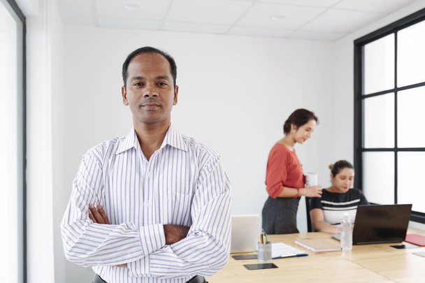 Portrait of Indian serious male leader standing with arms crossed and looking at camera with employees working on laptop in the background at office - Fotografie, Obrázek