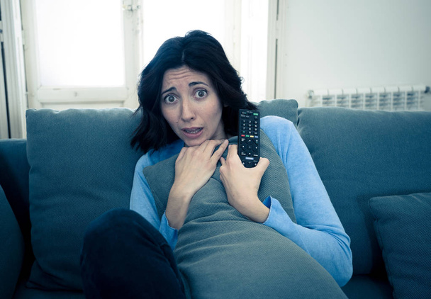 Lifestyle portrait of woman feeling scared and shocked making fear, anxiety gestures while watching horror movie on TV holding remote control. In horror and violence on TV and internet and mass media. - Foto, imagen