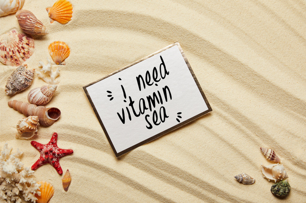 Draufsicht auf Karte mit Vitamin-Meer-Schriftzug in der Nähe von Muscheln, roten Seesternen und Korallen am Sandstrand  - Foto, Bild