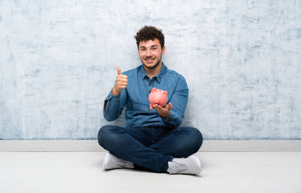 Mladý muž, který sedí na podlaze a drží velkou prasečí banku - Fotografie, Obrázek