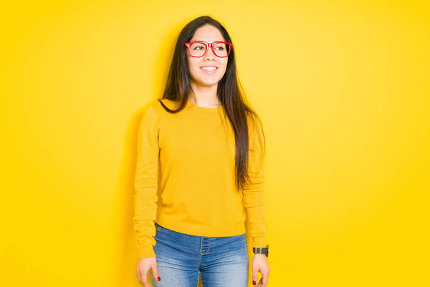 Belle femme brune portant des lunettes rouges sur fond jaune isolé regardant de côté avec le sourire sur le visage, expression naturelle. Rire confiant
. - Photo, image