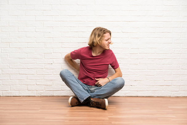 Hombre rubio sentado en el suelo sufriendo de dolor de espalda por haber hecho un esfuerzo
 - Foto, Imagen