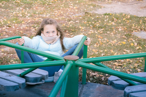 Mooi meisje gekleed in donsjack zittend op schommel op de speelplaats op koele herfst dag. - Foto, afbeelding