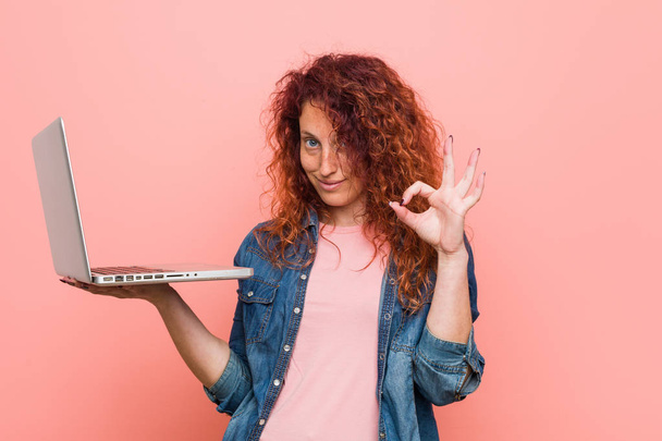 Young caucasian redhead woman holding a laptop cheerful and confident showing ok gesture. - Photo, Image