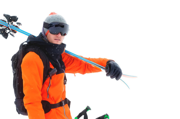 Portret van een professionele atleet skiër in een oranje jasje met een zwart masker en met ski's op zijn schouder kijkt in de camera. Geïsoleerd op wit - Foto, afbeelding