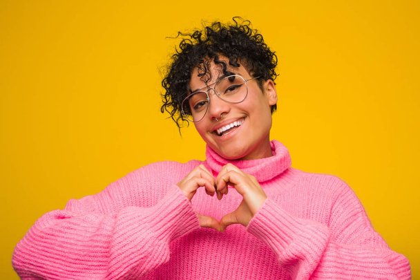 ピンクのセーターを着た若いアフリカ系アメリカ人女性が微笑み、手でハートの形を見せる. - 写真・画像