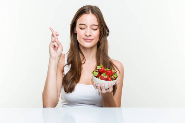 Young caucasian woman holding a strawberries bowl crossing fingers for having luck - Photo, Image