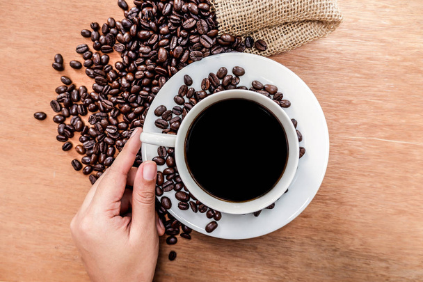 Tasse Kaffee mit gerösteten Bohnen und Hand auf Holzhintergrund, Draufsicht - Foto, Bild