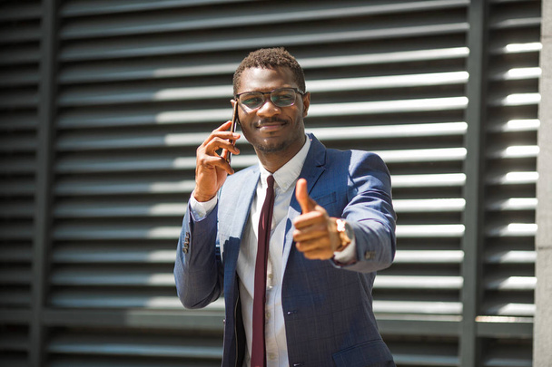 handsome young adult african man in suit and glasses talking on the phone with hand gesture - Foto, imagen