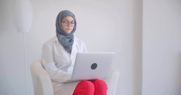 Closeup portrait of young muslim attractive female doctor in hijab and white coat using the laptop looking at camera smiling happily sitting in the armchair in the white room indoors - Záběry, video