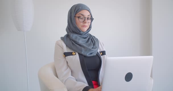 Closeup portrait of young muslim attractive female doctor in hijab and eyeglasses using the laptop looking at camera sitting in the armchair in the white room indoors - Materiał filmowy, wideo