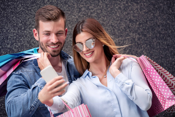 Pareja feliz con bolsas de compras tomando selfie con teléfono inteligente. - Foto, Imagen