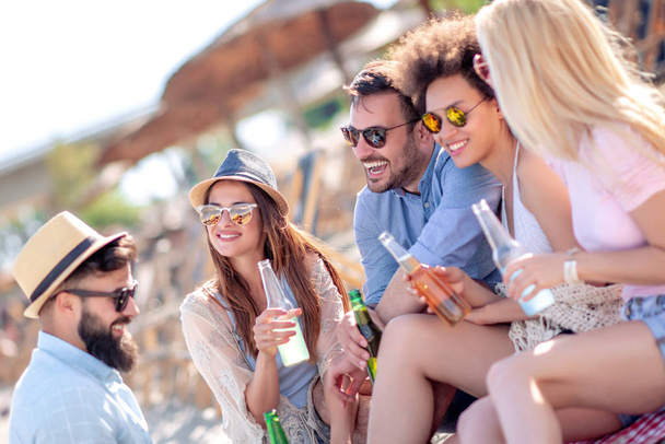 Grupa młodych przyjaciół o imprezie na plaży. Mężczyźni i kobiety piją piwo i bawią się razem. - Zdjęcie, obraz