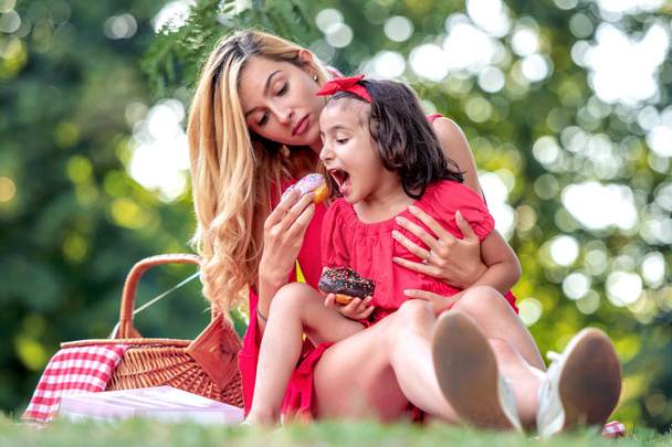 Young mother and her daughter have fun,eating a donut in the park. - Фото, зображення