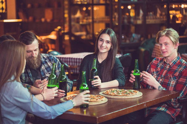 Friends having a drinks in a bar, They are sitting at a wooden table with beers and pizza - Fotoğraf, Görsel
