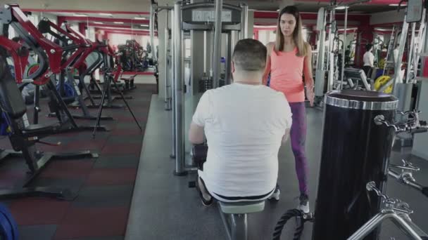 Man shakes his arm muscles on training simulator in gym with private coach. Weight loss drills from personal individual instructor for fat guy in fitness club. Thick person doing physical exercises - Filmmaterial, Video